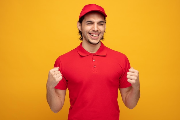joyful young delivery man wearing uniform and cap showing yes gesture with eyes closed isolated on yellow background