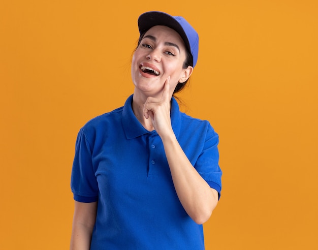 Радостная молодая женщина-доставщик в униформе и кепке, глядя на переднее трогательное лицо пальцем, изолированным на оранжевой стене с копией пространства