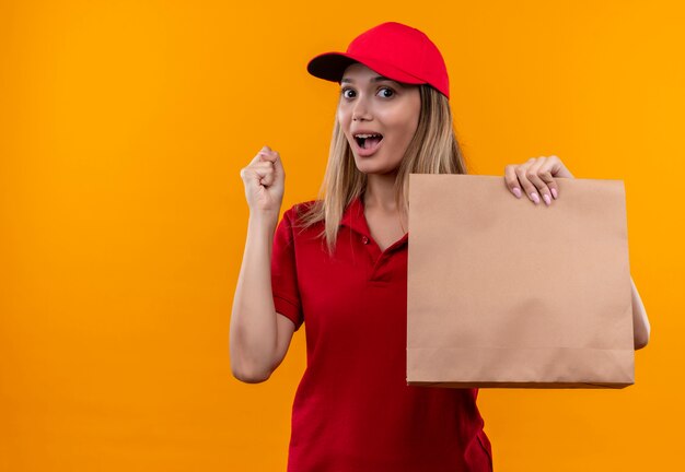 Радостная молодая доставщица в красной форме и кепке держит бумажный пакет и показывает жест "да"