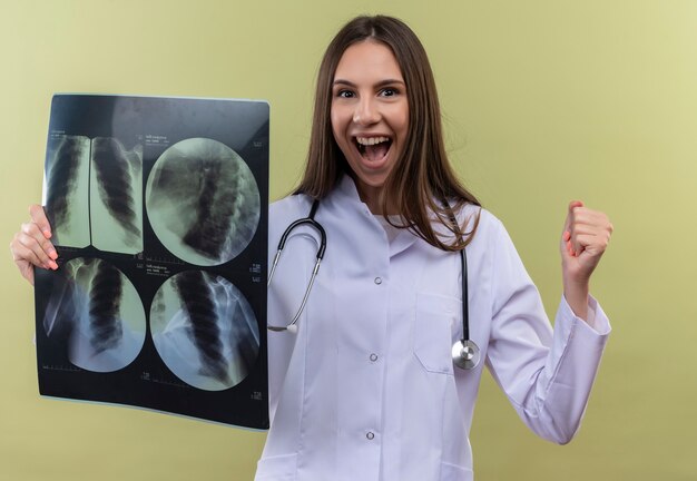 녹색 벽에 예 제스처를 보여주는 엑스레이를 들고 청진 기 의료 가운을 입고 즐거운 젊은 의사 소녀
