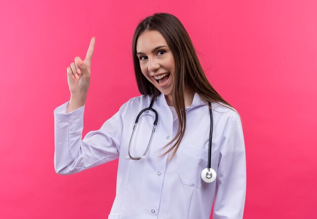 聴診器の医療用ガウンを身に着けているうれしそうな若い医者の女の子は、孤立したピンクの壁に指を指す