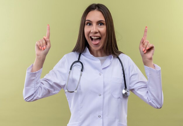 Радостная молодая девушка-врач в медицинском халате со стетоскопом показывает языком, указывающим на зеленую стену