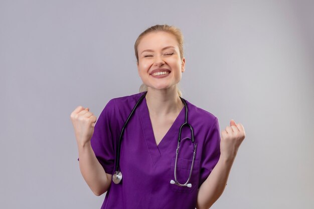 Joyful young doctor wearing purple medical gown and stethoscope doing strong gesture on isolated white wall