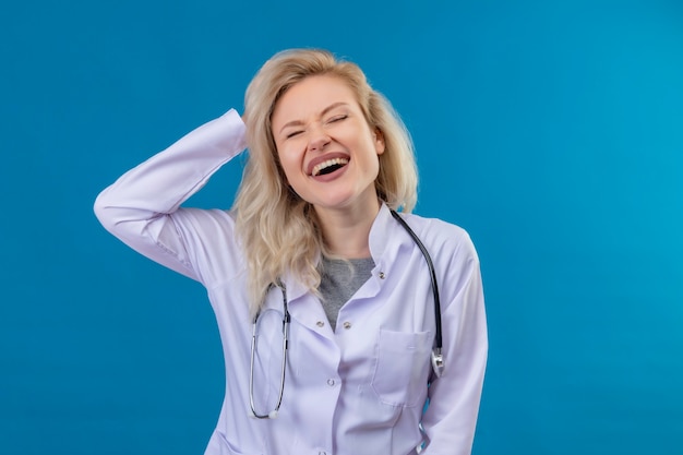 Joyful young doctor wearing stethoscope in medical gown grabbed head on blue wall