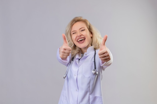 Foto gratuita gioioso giovane medico indossa uno stetoscopio in abito medico i suoi pollici in su sul muro bianco