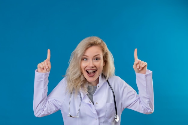 Joyful young doctor wearing stethoscope in medical gown points to up on blue wall