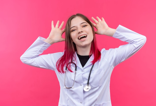 Радостная молодая женщина-врач в медицинском халате со стетоскопом положила руку на уши на розовой изолированной стене