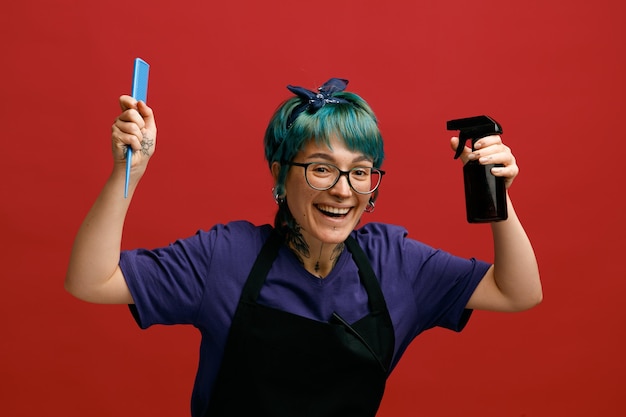 Joyful young female barber wearing uniform glasses and headband raising comb and hair spray up looking at camera isolated on red background