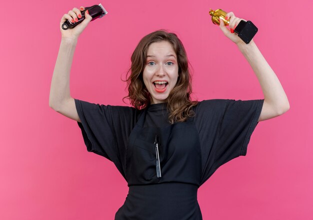 joyful young female barber wearing uniform raising up hair clippers and winner cup