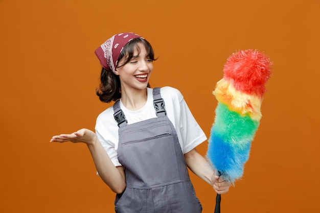 Gioiosa giovane donna addetta alle pulizie che indossa uniforme e bandana che tiene uno spolverino di piume guardandolo mostrando la mano vuota isolata su sfondo arancione