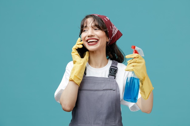 Joyful young female cleaner wearing uniform bandana and rubber gloves holding cleanser looking at side while talking on phone isolated on blue background
