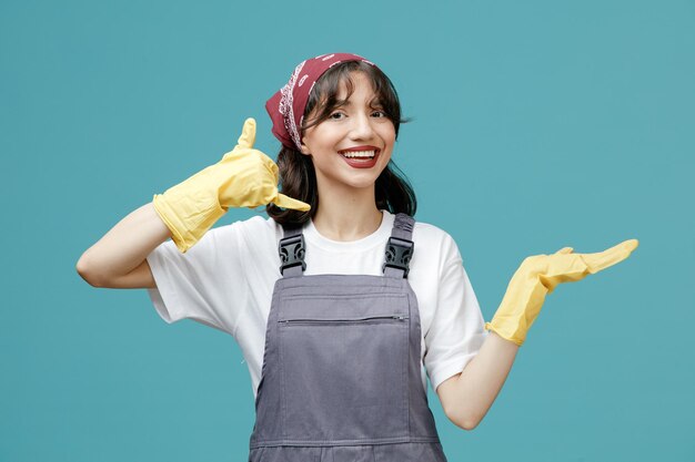 Joyful young female cleaner wearing uniform bandana and rubber gloves looking at camera showing empty hand making call gesture isolated on blue background