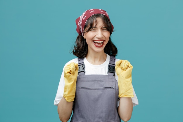 Joyful young female cleaner wearing uniform bandana and rubber gloves looking at camera showing yes gesture isolated on blue background