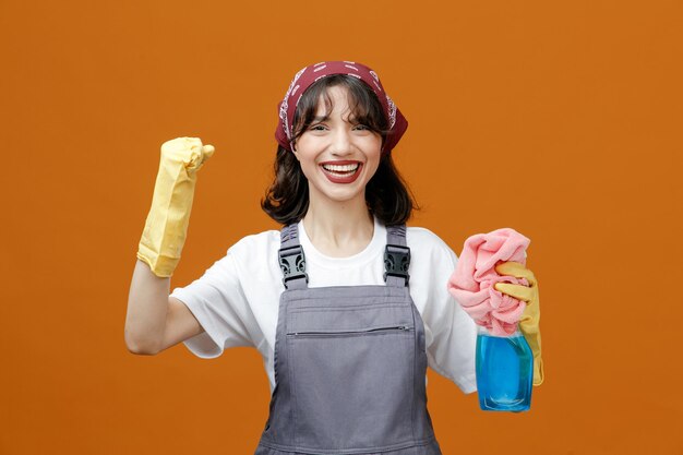 Joyful young female cleaner wearing uniform rubber gloves and bandana holding cloth duster and cleanser keeping fist in air looking at camera isolated on orange background