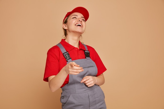 Joyful young female construction worker wearing uniform and cap looking up pointing at camera laughing 