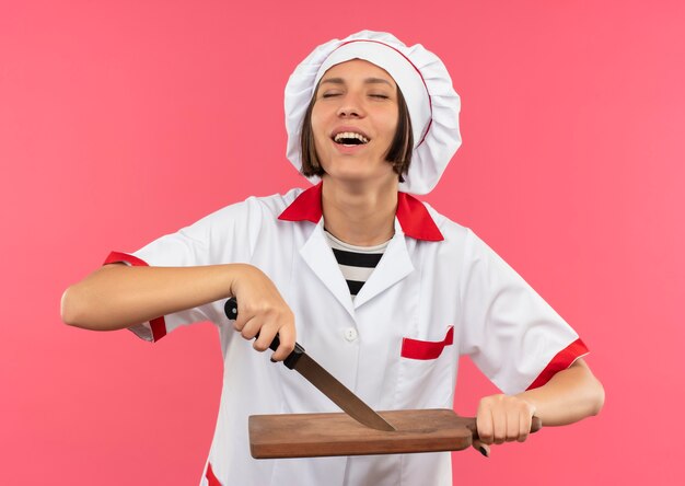 ピンクで隔離の目を閉じてナイフとまな板を保持しているシェフの制服を着たうれしそうな若い女性料理人