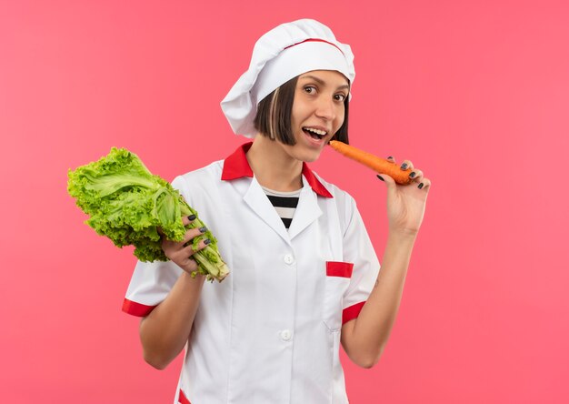 Joyful young female cook in chef uniform holding lettuce and trying to bite carrot isolated on pink