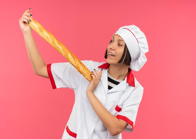 Joyful young female cook in chef uniform holding and looking at bread stick isolated on pink