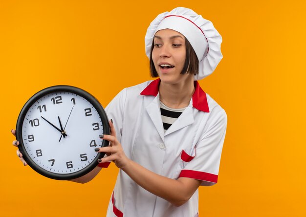 요리사 유니폼 잡고 오렌지에 고립 된 시계를보고 즐거운 젊은 여성 요리사