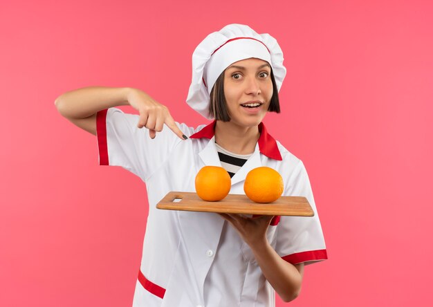 핑크에 고립 된 그것에 오렌지와 커팅 보드를 들고 요리사 유니폼에 즐거운 젊은 여성 요리사