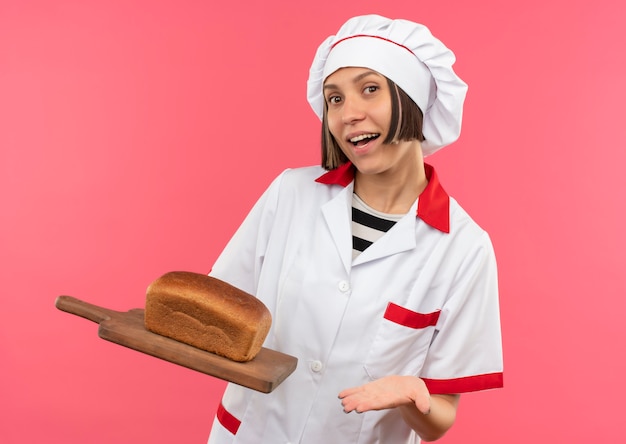 Gioioso giovane cuoco femminile in uniforme del cuoco unico che tiene e che indica con la mano al tagliere con pane su di esso isolato sul colore rosa