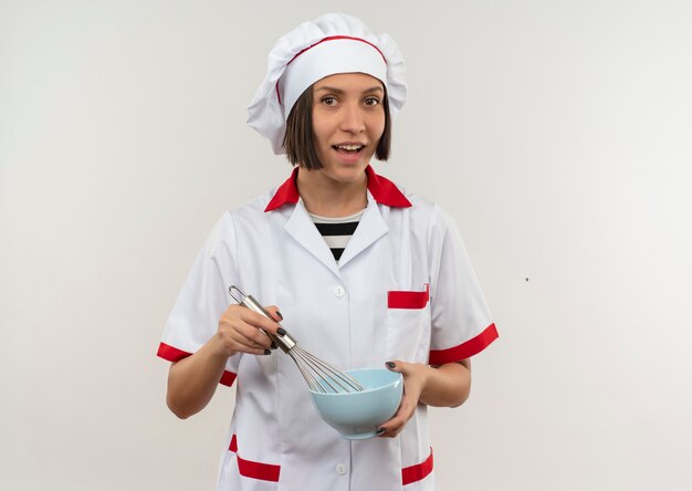 Joyful young female cook in chef uniform holding whisk and bowl isolated on white