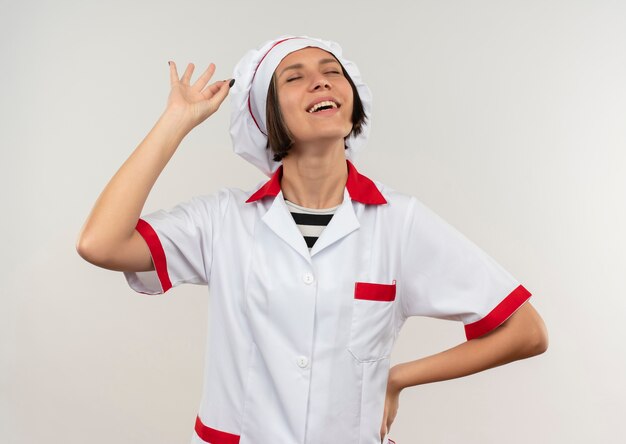 Joyful young female cook in chef uniform putting hand on waist doing ok sign with closed eyes isolated on white