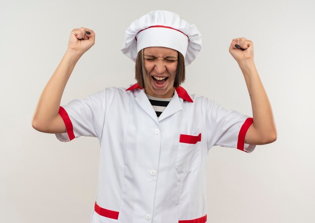 Joyful young female cook in chef uniform raising fists with closed eyes isolated on white