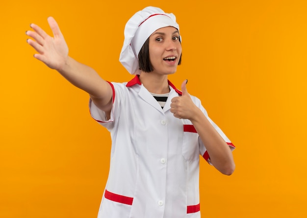 Joyful young female cook in chef uniform showing thumb up and stretching out hand at front isolated on orange