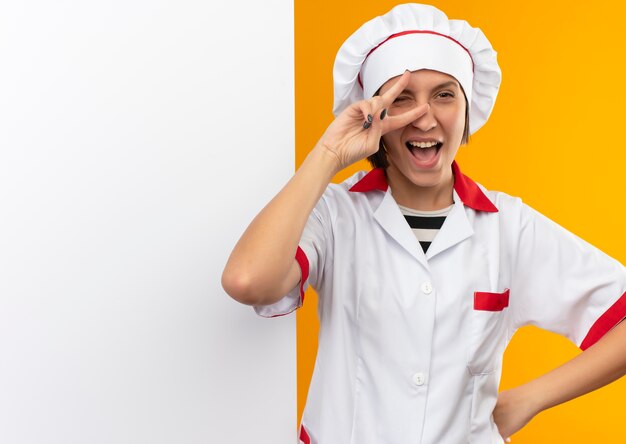 오렌지에 고립 된 평화 서명 하 고 허리에 손을 넣어 흰 벽 앞에 서있는 요리사 유니폼에 즐거운 젊은 여성 요리사