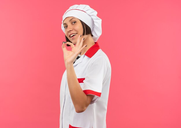 Joyful young female cook in chef uniform standing in profile view doing ok sign isolated on pink