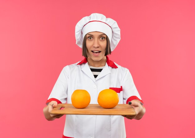 Joyful young female cook in chef uniform stretching out cutting board with oranges on it isolated on pink