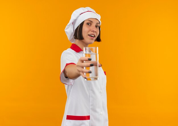 Joyful young female cook in chef uniform stretching out glass of water isolated on orange
