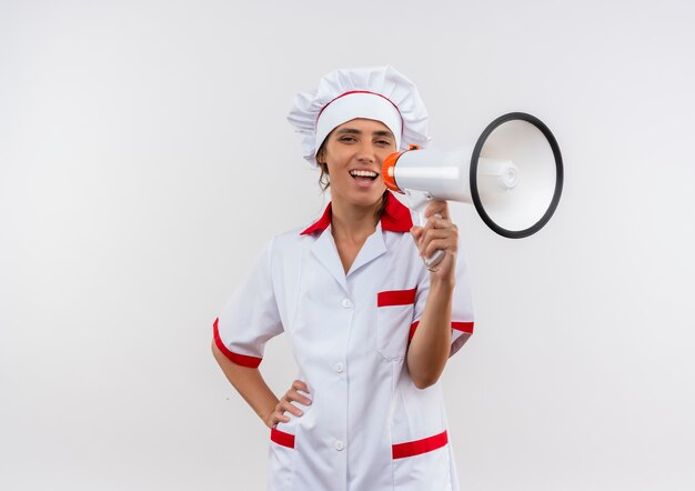 Joyful young female cook wearing chef uniform speake on loudspeaker putting hand on hip on isolated white wall with copy space