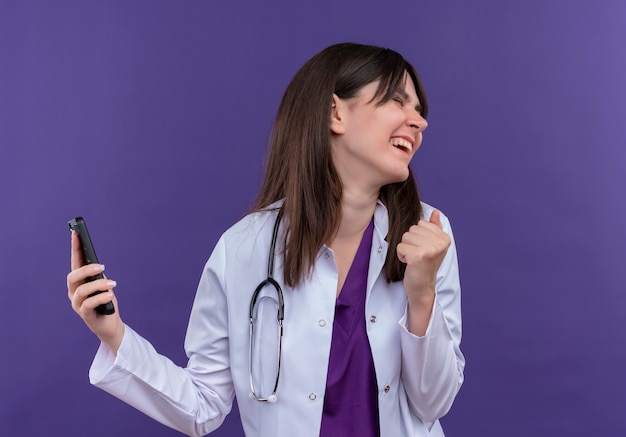 Joyful young female doctor in medical robe with stethoscope holds phone and looks to the side on isolated violet background with copy space