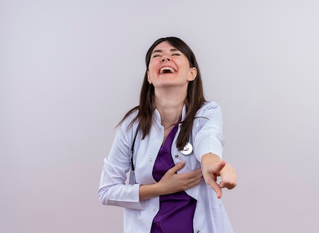 Joyful young female doctor in medical robe with stethoscope points at camera on isolated white background with copy space