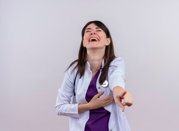 Радостная молодая женщина-врач в медицинском халате со стетоскопом указывает на камеру на изолированном белом фоне с копией пространства