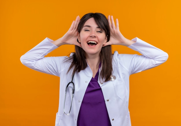 Joyful young female doctor in medical robe with stethoscope puts hands on her head on isolated orange background