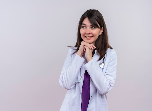 Joyful young female doctor in medical robe with stethoscope puts hands together on chin and looks to the side on isolated orange background with copy space