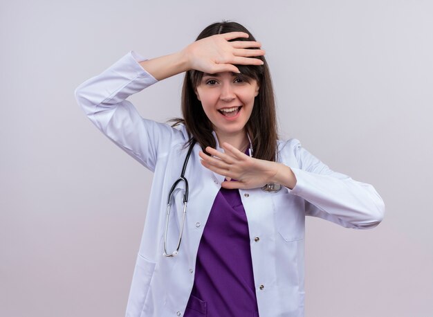 Joyful young female doctor in medical robe with stethoscope raises hands up on isolated white background