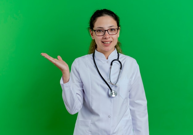 Радостная молодая женщина-врач в медицинском халате, стетоскопе и очках показывает пустую руку, изолированную на зеленой стене с копией пространства