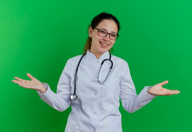 緑の壁に隔離された空の手を示す医療ローブと聴診器と眼鏡を身に着けているうれしそうな若い女性医師