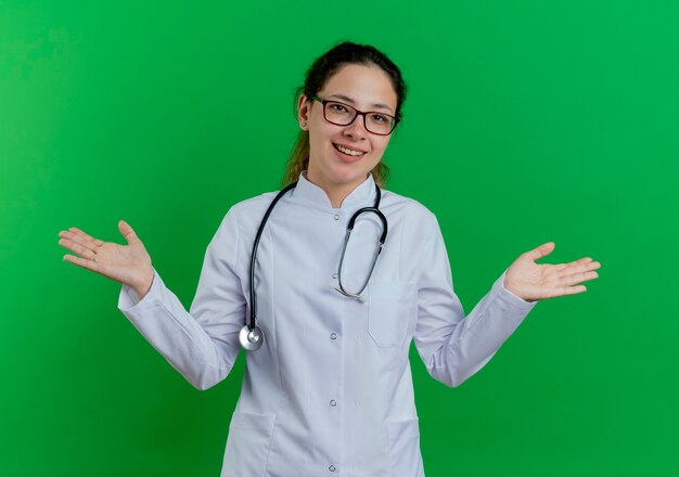 Радостная молодая женщина-врач в медицинском халате и стетоскопе и очках показывает пустые руки, изолированные на зеленой стене