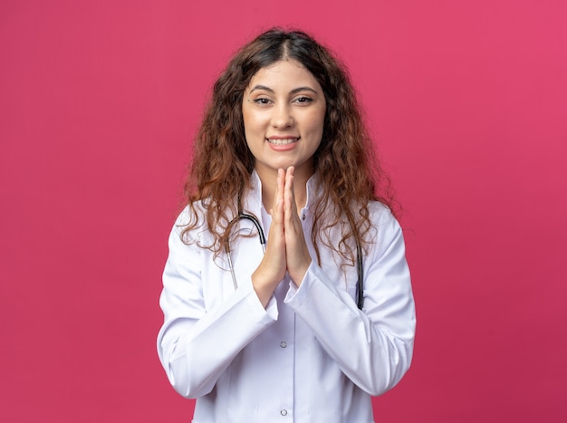 Радостная молодая женщина-врач в медицинском халате и стетоскопе держит руки в жесте молитвы, глядя на переднюю часть, изолированную на розовой стене с копией пространства