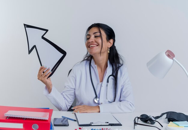 의료 가운과 청진기를 착용하고 의료 도구를 들고 고립 된 측면을 가리키는 화살표 표시를보고 책상에 앉아 즐거운 젊은 여성 의사