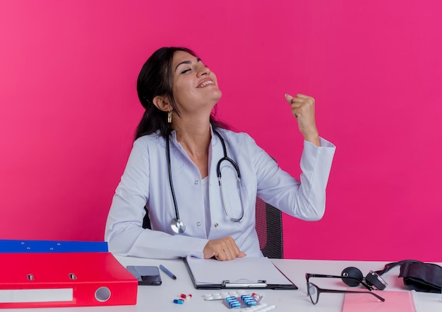 Joyful young female doctor wearing medical robe and stethoscope sitting at desk with medical tools putting fist on desk doing yes gesture with closed eyes isolated on pink wall