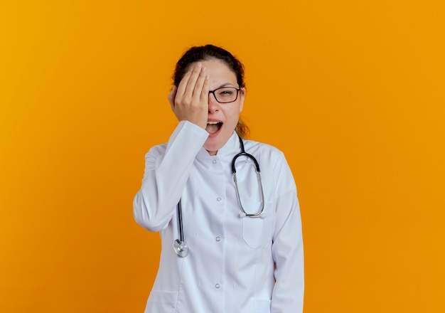 Joyful young female doctor wearing medical robe and stethoscope with glasses covered with hand eye isolated