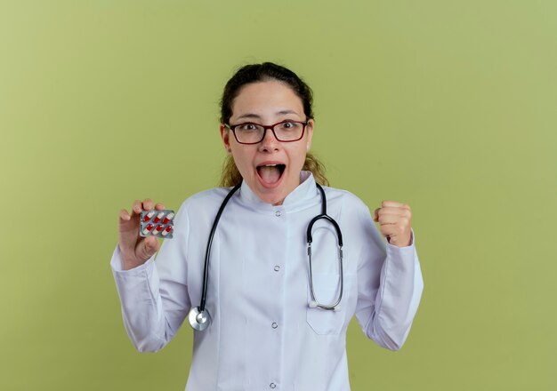 Радостная молодая женщина-врач в медицинском халате и стетоскопе в очках держит таблетки и показывает жест да изолированы