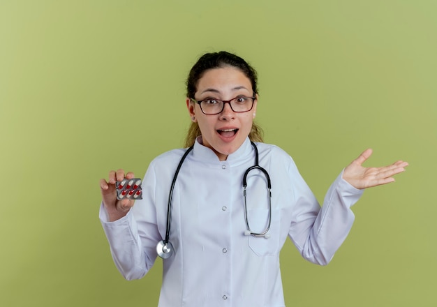 Радостная молодая женщина-врач в медицинском халате и стетоскопе в очках держит таблетки и протягивает руку, изолированную на оливково-зеленой стене