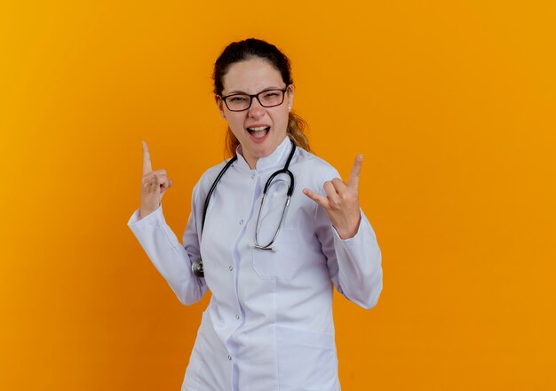 Joyful young female doctor wearing medical robe and stethoscope with glasses showing goat gestures isolated